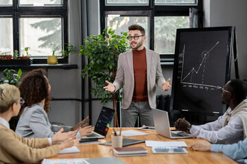 Poster - Confident business coach explaining graph by blackboard to young intercultural managers