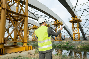 Poster - Rear view of engineer with blueprint looking at construction building and talking on mobile phone outdoors