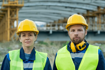 Poster - Portrait of two engineers in work helmets looking at camera while working together on construction site