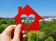 Hand Holding Red Felt House On Blurred Background Of Cottage Village In Summer Sunny Day. Heart-shaped Hole Is Cut Instead Of A Window. Buy Or Build Your Dream Home, Real Estate Agency Concept.