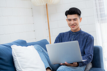 Young asian businessman smile and work from home with laptop computer online to internet on sofa in living room, freelance man using notebook on couch with comfort, new normal, lifestyle concept.