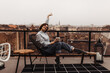 © Look! - Happy woman in jeans, blue shirt and cool socks rises arm, sits on chair holds coffee cup, smiles on terrace with city view.