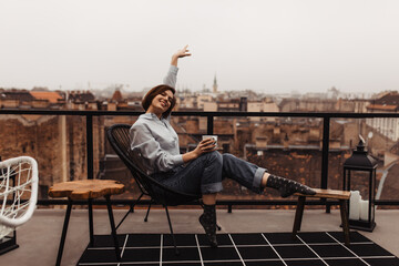 Happy woman in jeans, blue shirt and cool socks rises arm, sits on chair holds coffee cup, smiles on terrace with city view.