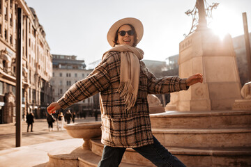 Wall Mural - Stylish young woman in checkered coat and hat smiles sincerely on city square. Beautiful girl in sunglasses explores European town.