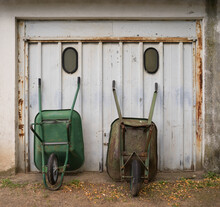 Two Old Wheelbarrows Stand Upright In Front Of A Garage Door