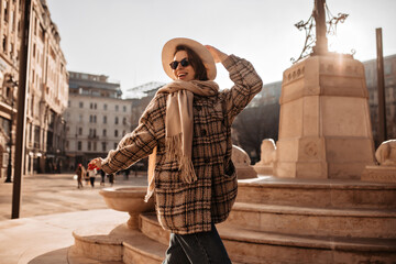 Wall Mural - Attractive good-humored girl holds beige hat and explores European city. Pretty woman in checkered coat smiles and walks near statue on town square.