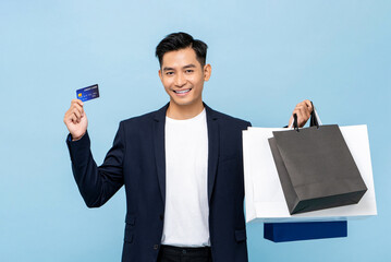 Wall Mural - Young handsome southeast Asian millenial man holding credit card and bags in light blue studio isolated background for cashless shopping concept