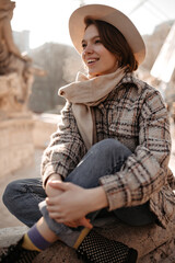 Wall Mural - Pretty short-haired woman in stylish checkered coat and jeans smiles widely outside. Young lady sits near fountain.