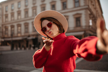 Wall Mural - Cool woman in beige hat blows kiss and takes selfie outside. Charming girl in red sweater walks outdoors.