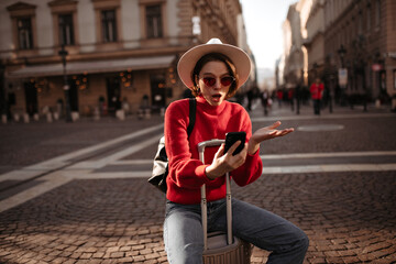 Shocked woman in red sweater and jeans sits on suitcase outside. Surprised girl in hat reads message and holds phone.