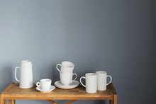 White Cups And Jug On Wooden Shelf On Gray Background