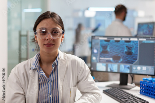 Scientist with lab coat sitting in laboratory looking at camera smiling, while colleague working on computer in background. Chemist examining virus evolution using high tech for scientific research