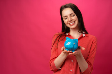 Happy smiling young woman holding blue piggy bank against pink background