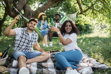 Wall Mural - Indian man and african woman relaxing on played on grass with beer in hands while another couple sitting in hammock and chatting on background. Happy friends enjoying picnic outdoors.