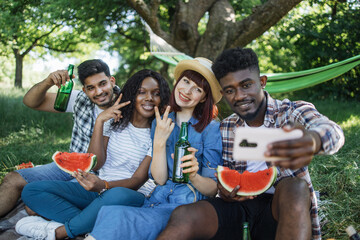 Wall Mural - Cheerful group of four diverse friends sitting at green garden with watermelon and beer in hands and taking selfie on modern smartphone, Concept of people, technology and leisure time.