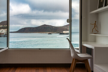 Atlantic ocean and landscape view through open window from inside home or hotel room with working table and chair. Work remote from home office in beach apartment. Gran Canaria, Canary Islands, Spain.