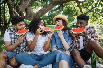 Wall Mural - Joyful men and women sitting together at green garden and eating sweet watermelon. Group of four friends having picnic in nature. Concept of summer time and vacation.