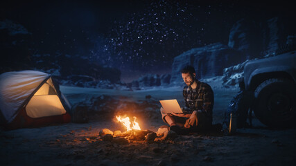 Night Tent Camping in Canyon: Male Traveler Uses Laptop Computer Sitting by Campfire. Man on Digital Remote Work, e-shopping, ecommerce, Using Internet, Social Media Posting on Vacation Trip 