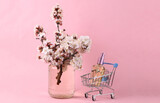 Fototapeta Mapy - Shopping trolley with perfume bottle and Beautiful flowering branches in glass jar on pink background.