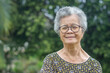 © meeboonstudio - Portrait of a senior woman with short gray hair, wearing glasses smiling, and looking at the camera while standing in a garden. Aged people and relaxation concept