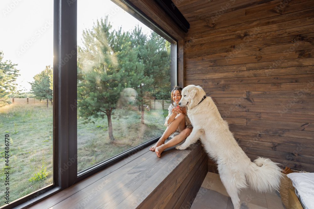 Young woman resting at beautiful country house or hotel, sitting on the window sill with pine forest view and hugs with big white dog. Concept of solitude and recreation on nature with pet - obrazy, fototapety, plakaty 