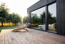Young Woman Resting On Sunbed And Reading On A Tablet On The Wooden Terrace Near The Modern House With Panoramic Windows Near Pine Forest. Concept Of Solitude And Recreation On Nature