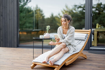 young woman resting on sunbed and looks on the phone on terrace at modern house with panoramic windo