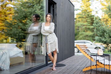 Young woman enjoy staying near panoramic window with pine forest view on terrace of wooden modern house. Concept of solitude and recreation on nature. Beautiful place for vacation.