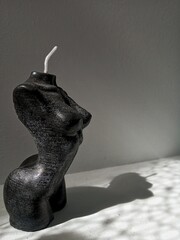 Black candle in the shape of a female body sideways, shade on white wall background.