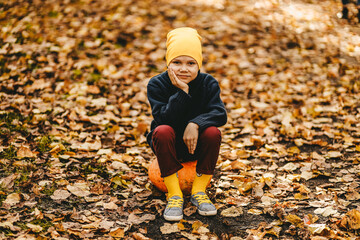 A small teenage boy in a yellow hat and warm clothes is walking and having fun in an autumn park in nature, a child is holding a pumpkin in his hands, a selective focus