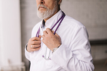 Wall Mural - Close-up cropped portrait of confident mature male doctor wearing white lab coat and stethoscope standing in light hospital office room, blurred background. Professional physician working at workplace