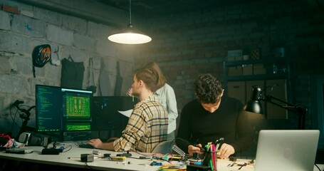 Electronics Workshop in Garage Office. Team Works in Technology Startup in Loft Basement or Attic at Night with Fog. IT Concept with Soldering and Coding. Leader Helps Coworker. 4K Medium Pan Video
