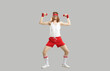 Full body skinny nerd with funny face doing exercise with dumbbells isolated on gray background. Full length hilarious smiling man in sweatband, white tank top and red shorts enjoying sports workout