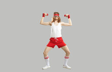 Full Body Skinny Nerd With Funny Face Doing Exercise With Dumbbells Isolated On Gray Background. Full Length Hilarious Smiling Man In Sweatband, White Tank Top And Red Shorts Enjoying Sports Workout