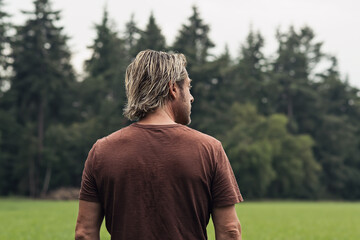 Wall Mural - Blonde man in a brown t-shirt stands in a field near a pine fore