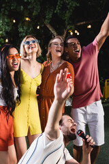 Wall Mural - Interracial friends with plastic cup singing near man with microphone during party outdoors