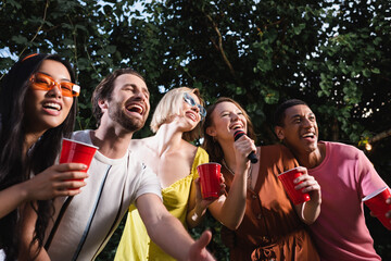 Wall Mural - Cheerful multiethnic people singing karaoke and holding drink in plastic cups outdoors