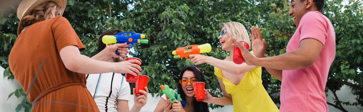 excited women playing with water guns during summer party with multiethnic friends, banner