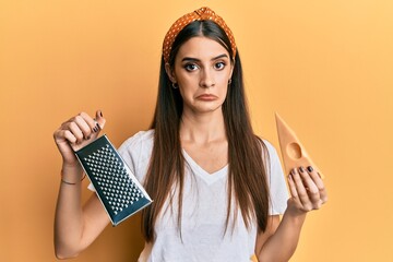 Beautiful brunette young woman holding grater and cheese depressed and worry for distress, crying angry and afraid. sad expression.