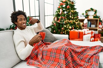 Sticker - Young african american man sitting on the sofa drinking coffee by christmas tree smiling cheerful showing and pointing with fingers teeth and mouth. dental health concept.