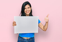 Young Hispanic Woman Holding Blank Empty Banner Smiling Happy And Positive, Thumb Up Doing Excellent And Approval Sign