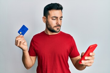 Canvas Print - Young hispanic man holding smartphone and credit card relaxed with serious expression on face. simple and natural looking at the camera.