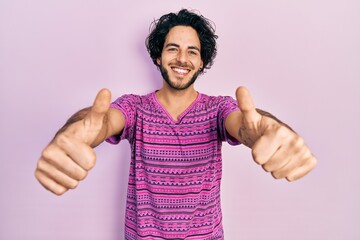 Handsome hispanic man wearing casual pink t shirt approving doing positive gesture with hand, thumbs up smiling and happy for success. winner gesture.