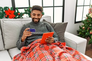 Wall Mural - Young african american man smiling happy using laptop and credit card sitting on the sofa at home.