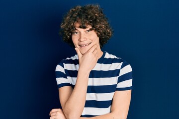 Handsome young man wearing casual striped t shirt looking confident at the camera smiling with crossed arms and hand raised on chin. thinking positive.