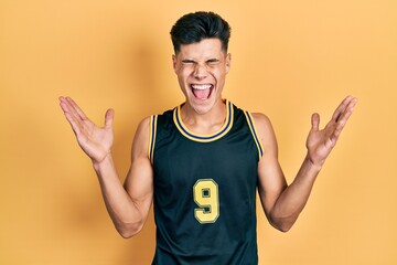 Wall Mural - Young hispanic man wearing basketball uniform celebrating mad and crazy for success with arms raised and closed eyes screaming excited. winner concept