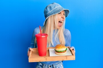 Poster - Young blonde woman eating a tasty classic burger and soda angry and mad screaming frustrated and furious, shouting with anger. rage and aggressive concept.