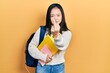 Young chinese girl holding student backpack and books showing middle finger, impolite and rude fuck off expression