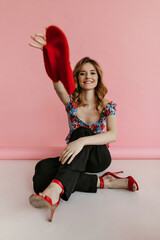 Wall Mural - Cool wavy haired woman in blue floral top and dark pants smiling. Stylish girl throws red beret on isolated pink background..