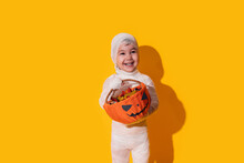 Child In Mummy Costume Holding Basket Of Chocolates In Front Of Yellow Background